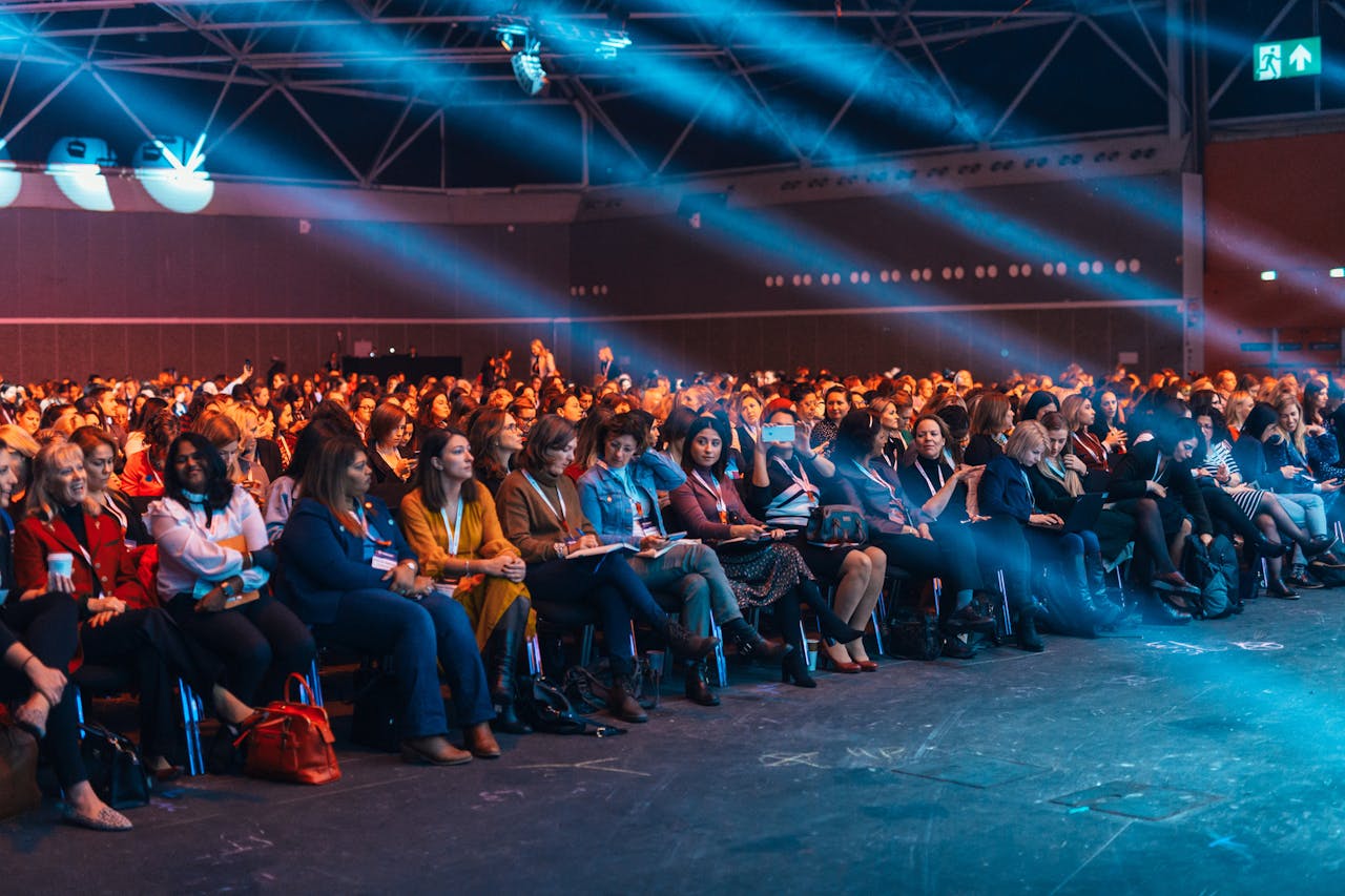 Home Vibrant image of a diverse audience seated in a large auditorium, highlighted by dynamic stage lighting.